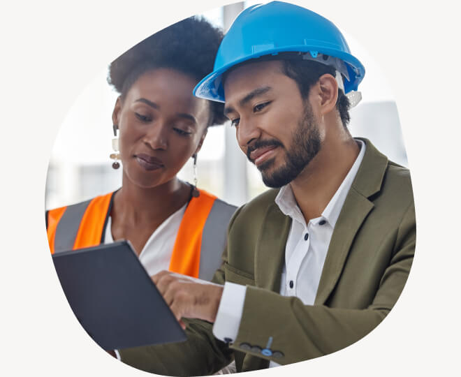 A woman wearing bright colored vest and a man with a hard hat and sport coat looking at a computer tablet. they appear to be at a construction site